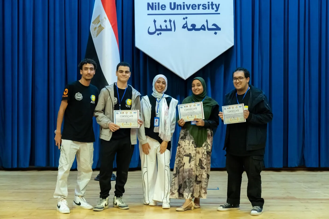 Five students holding certificates on stage under a Nile University banner