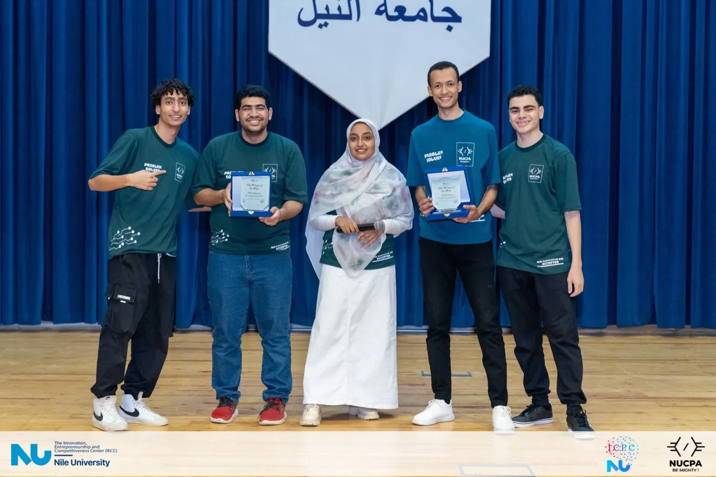 Four students in green contest shirts holding certificates on stage