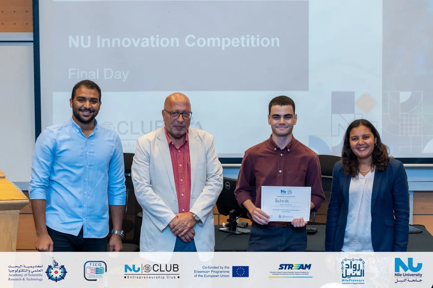 Four people on stage, one holding a certificate, in front of a NU Innovation Competition slide