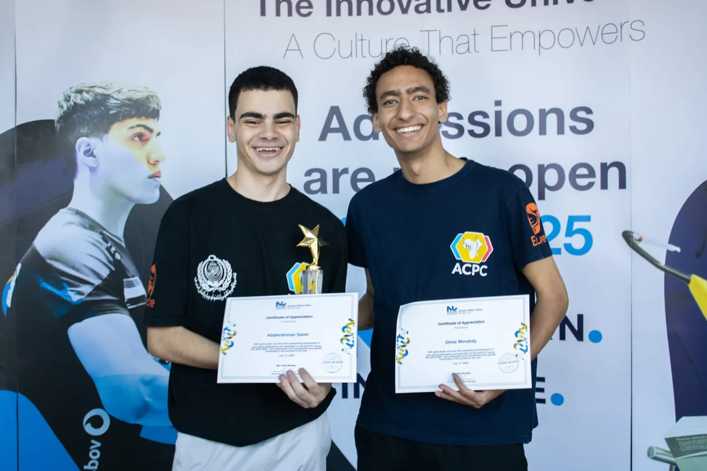 Two students holding certificates in front of a university banner